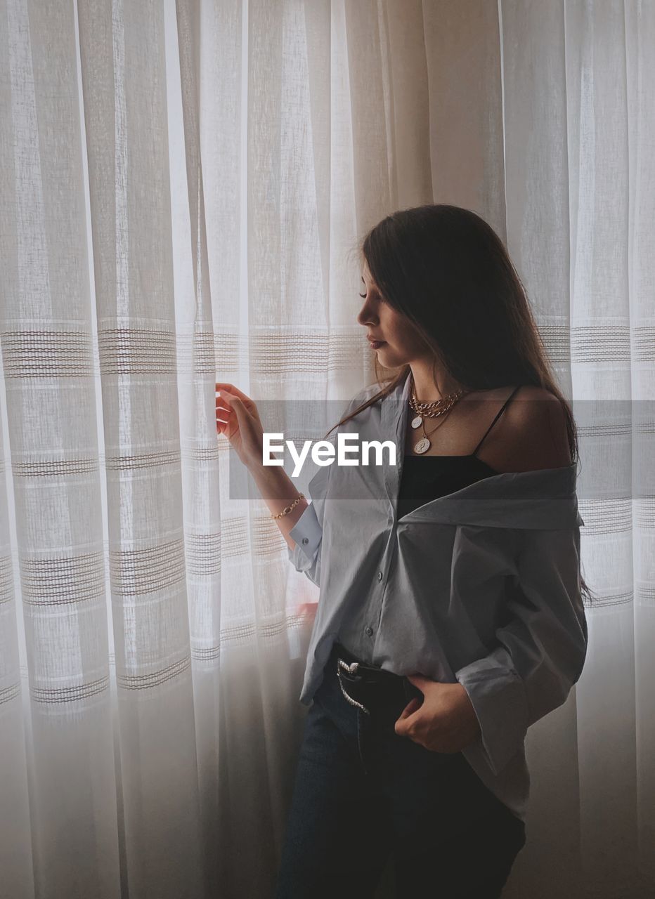 Woman looking away while standing against window at home