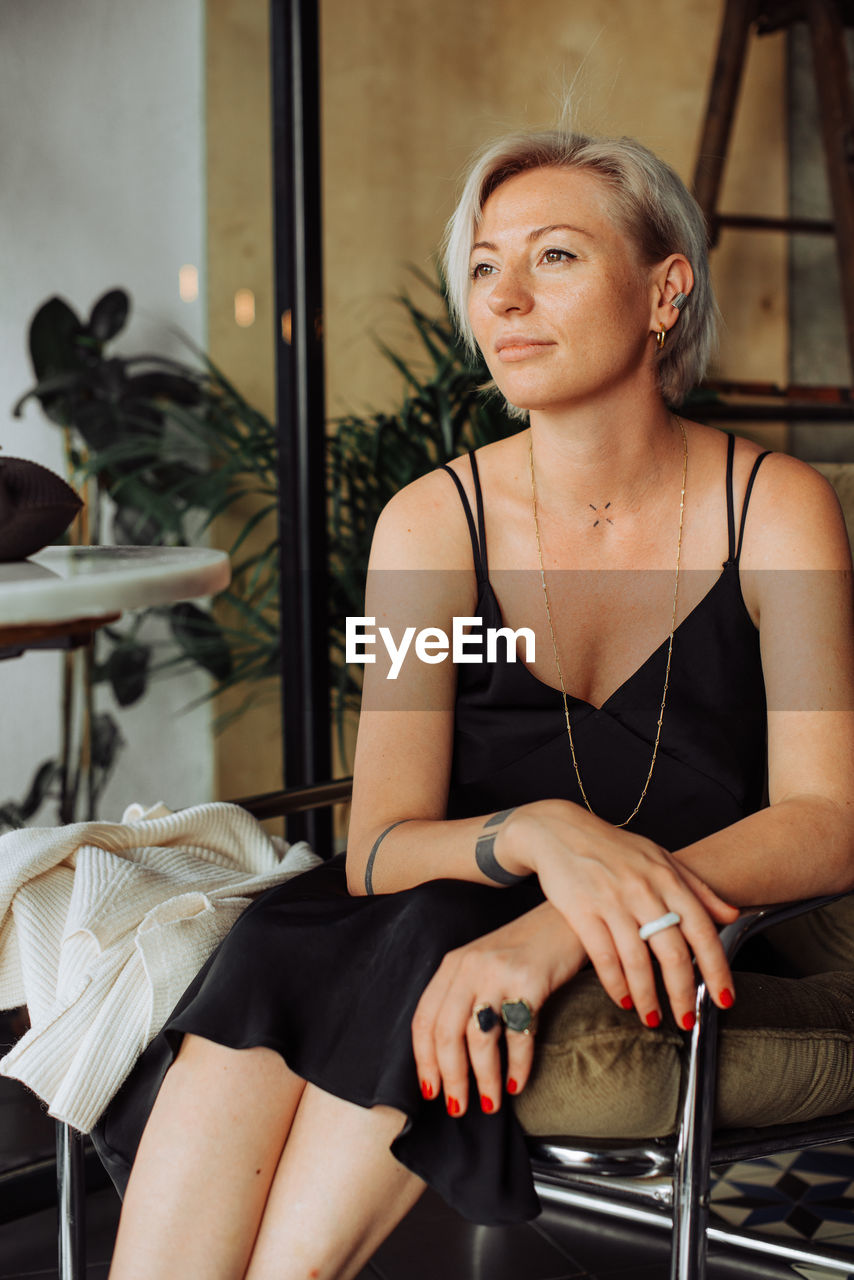 Blond stylish woman in black dress sitting in arm chair looking away