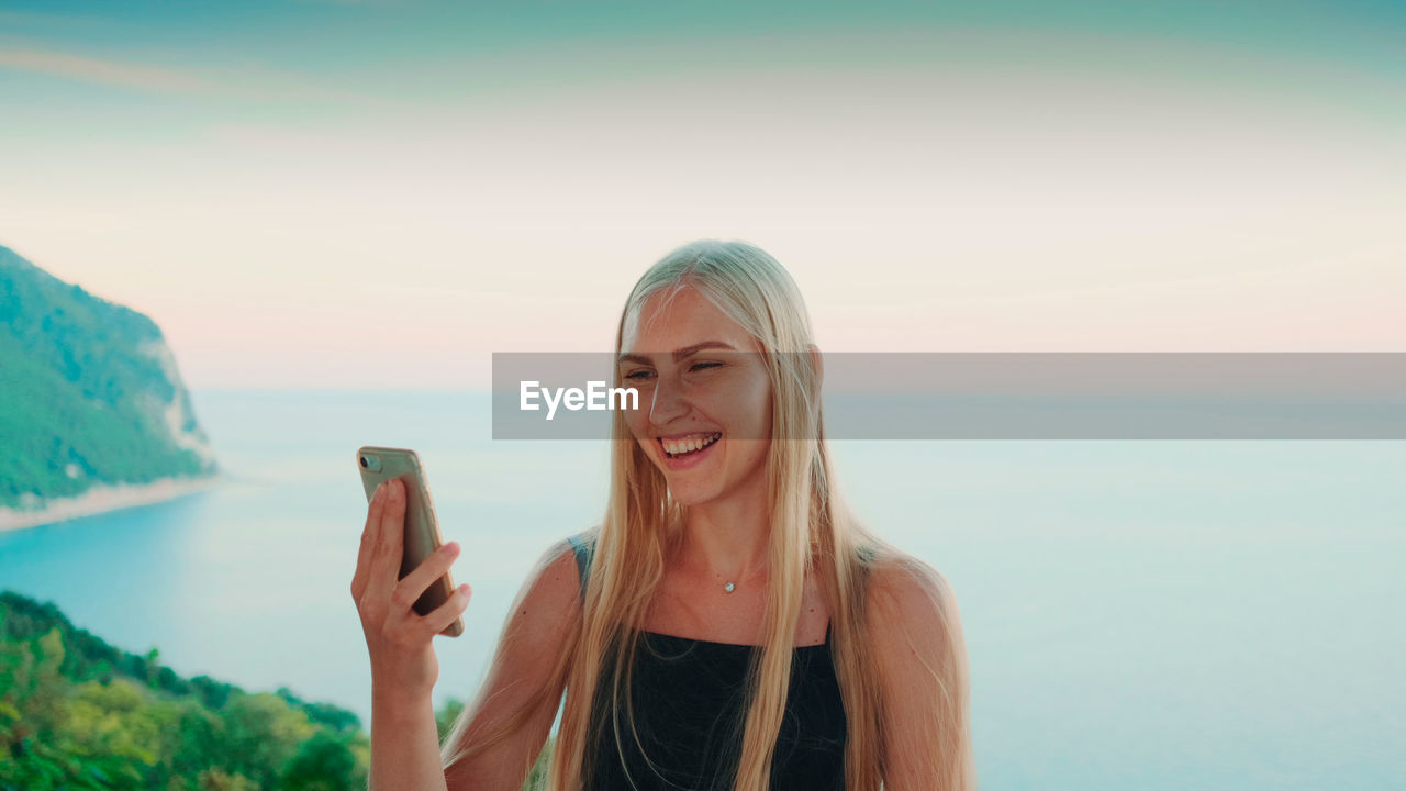 Smiling young woman using smart phone against sea