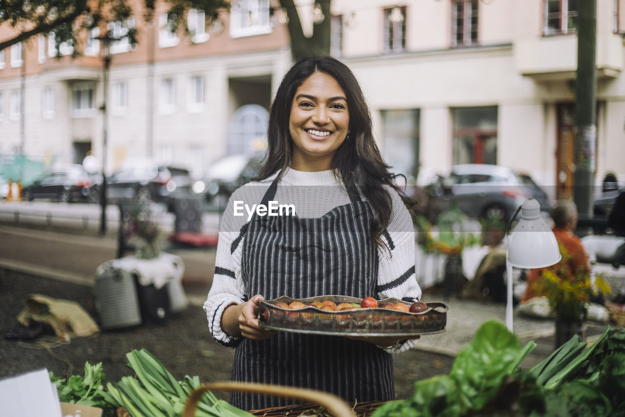 Portrait of smiling vendor holding vegetable tray while standing at farmer's market