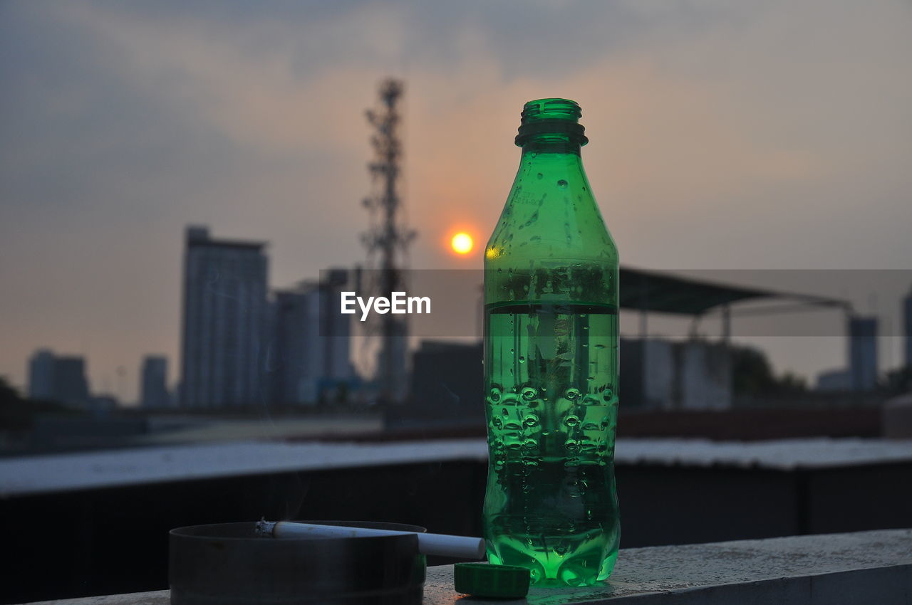 CLOSE-UP OF GLASS BOTTLES AGAINST CITY DURING SUNSET