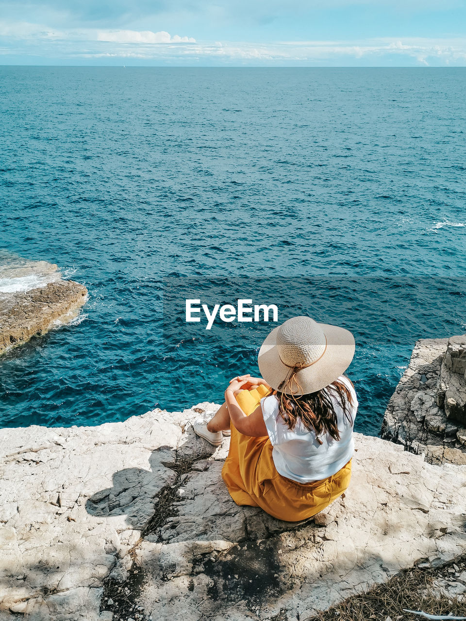 Young woman in yellow skirt sitting on edge of cliff above sea.