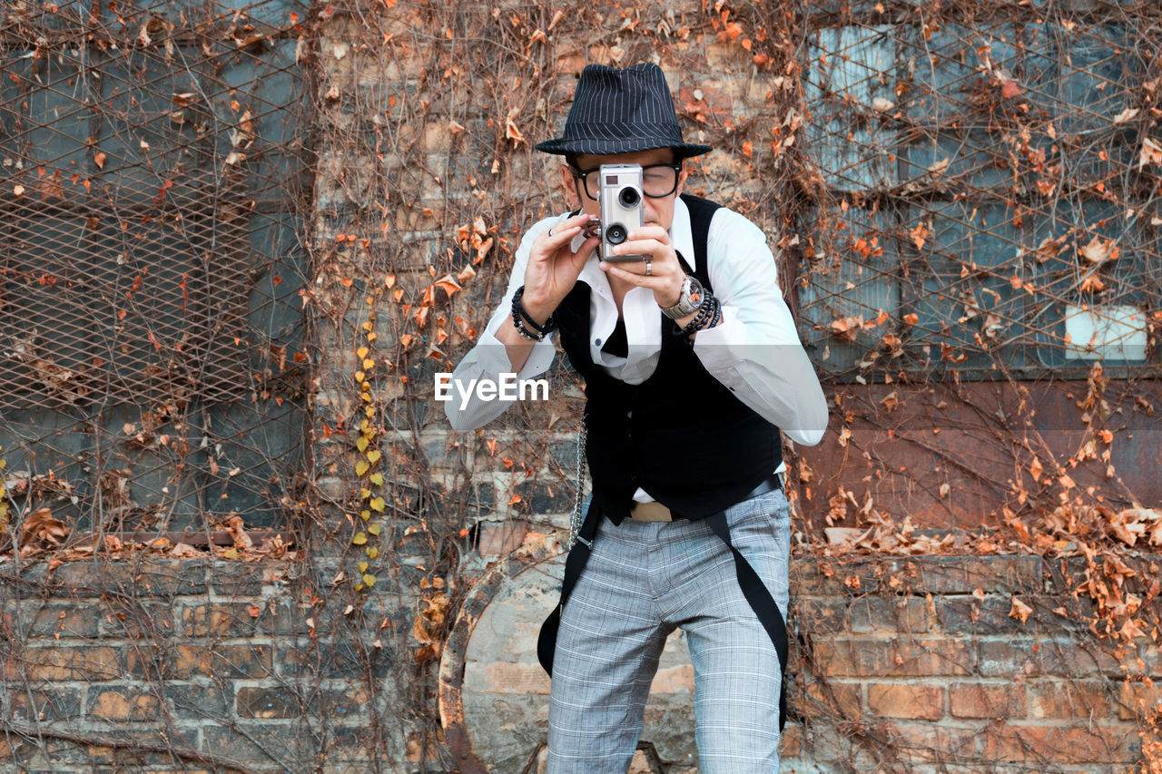 Mature man photographing while standing against brick wall during autumn