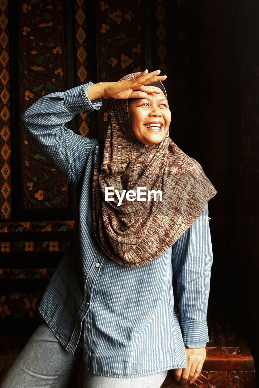 Cheerful woman wearing hijab while shielding eyes at home