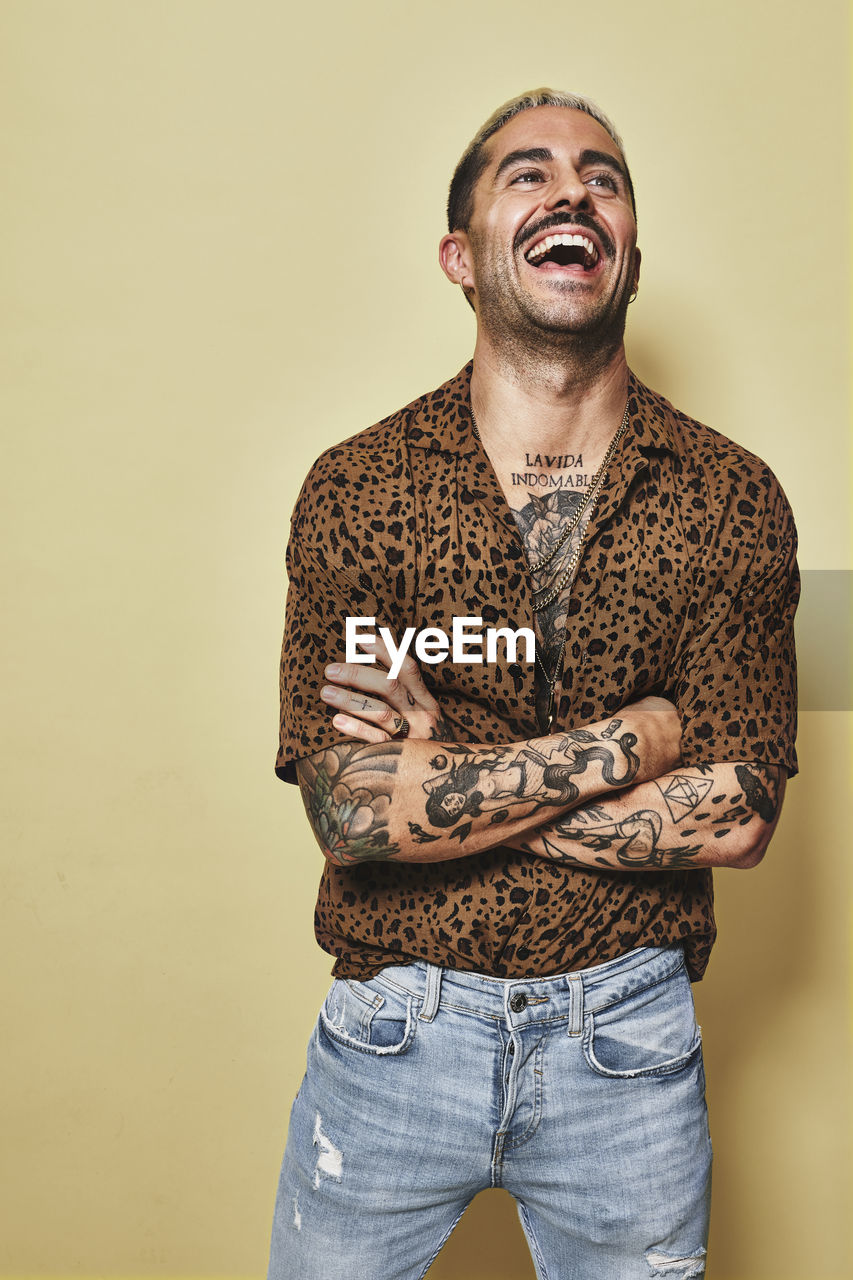 Cheerful fashionable male model with tattoos wearing trendy leopard shirt and jeans standing against beige background and looking away