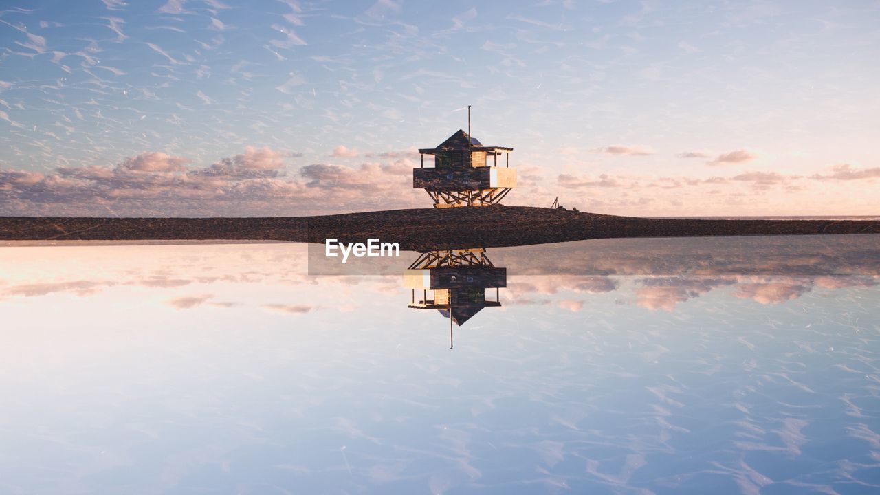 Reflection of lifeguard hut in sea at beach against sky during sunset