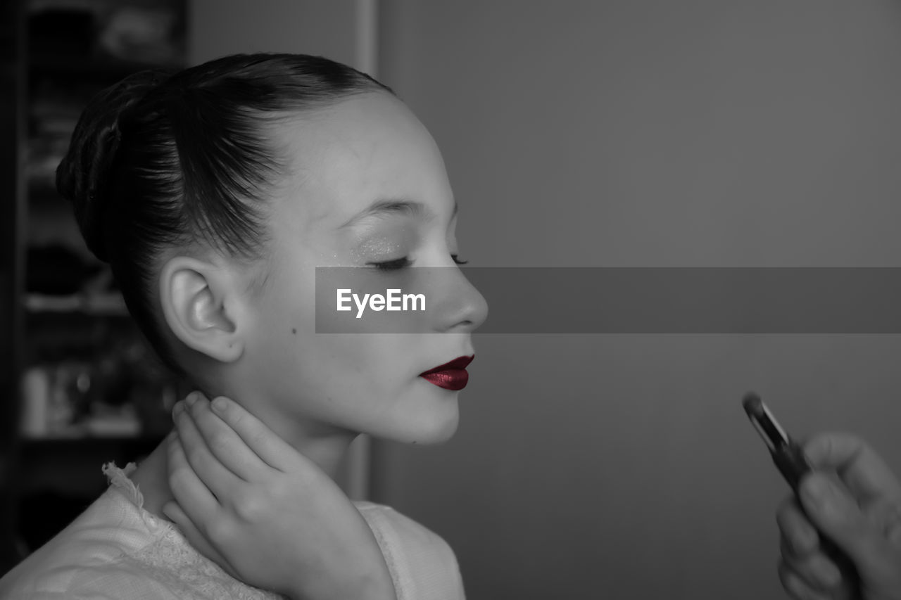 Close-up of girl with eyes closed applying red lipstick against wall
