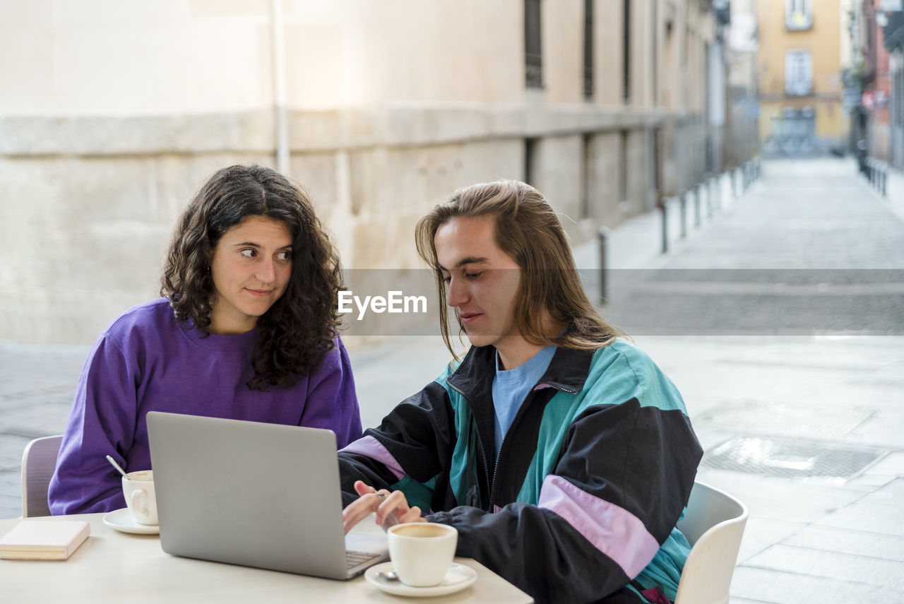 Two people using laptop and mobile phone while sitting outdoors at a coffee shop