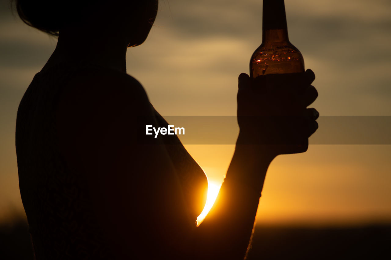 Silhouette woman holding bottle against sky during sunset