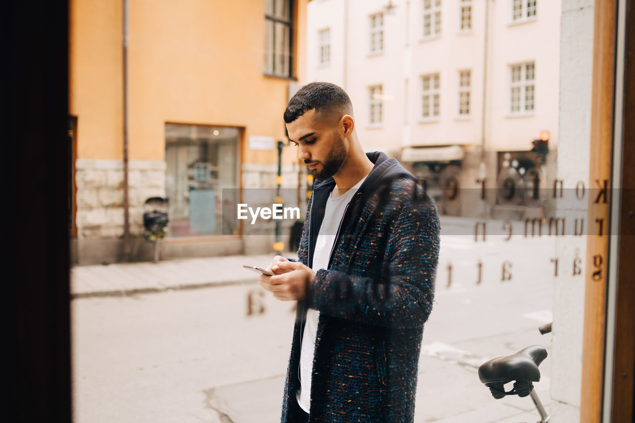 Confident young businessman using smart phone seen through window