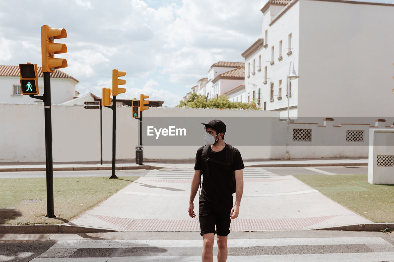 Unrecognizable guy in black outfit in respiratory mask strolling on street pavement under cloudy sky while looking away during covid 19 pandemic