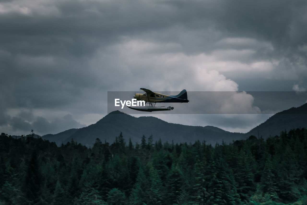 Low angle view of airplane flying over forest against cloudy sky