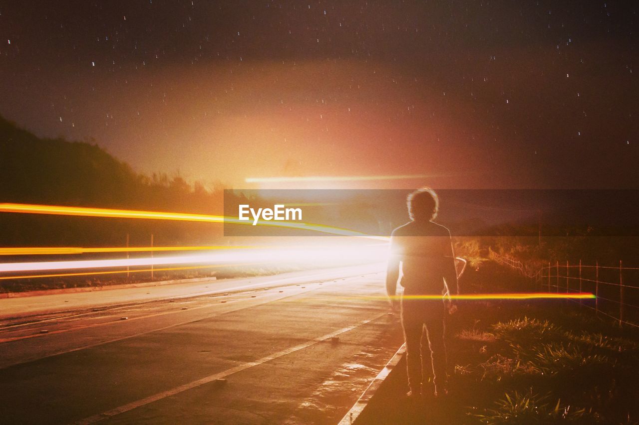 Rear view full length of man standing by light trails at roadside during night