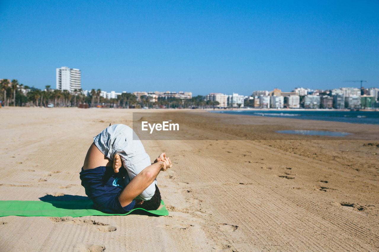 Full length of man performing yoga at beach by city against clear blue sky