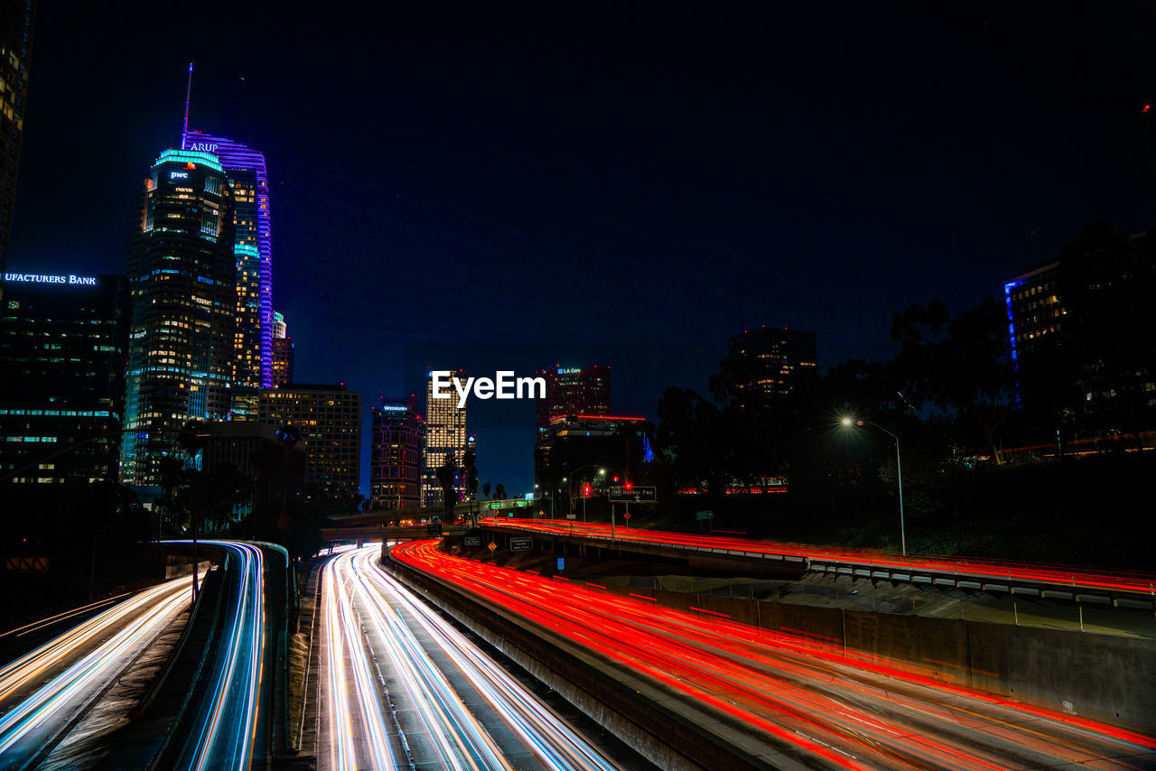 Light trails on road by illuminated buildings against sky at night