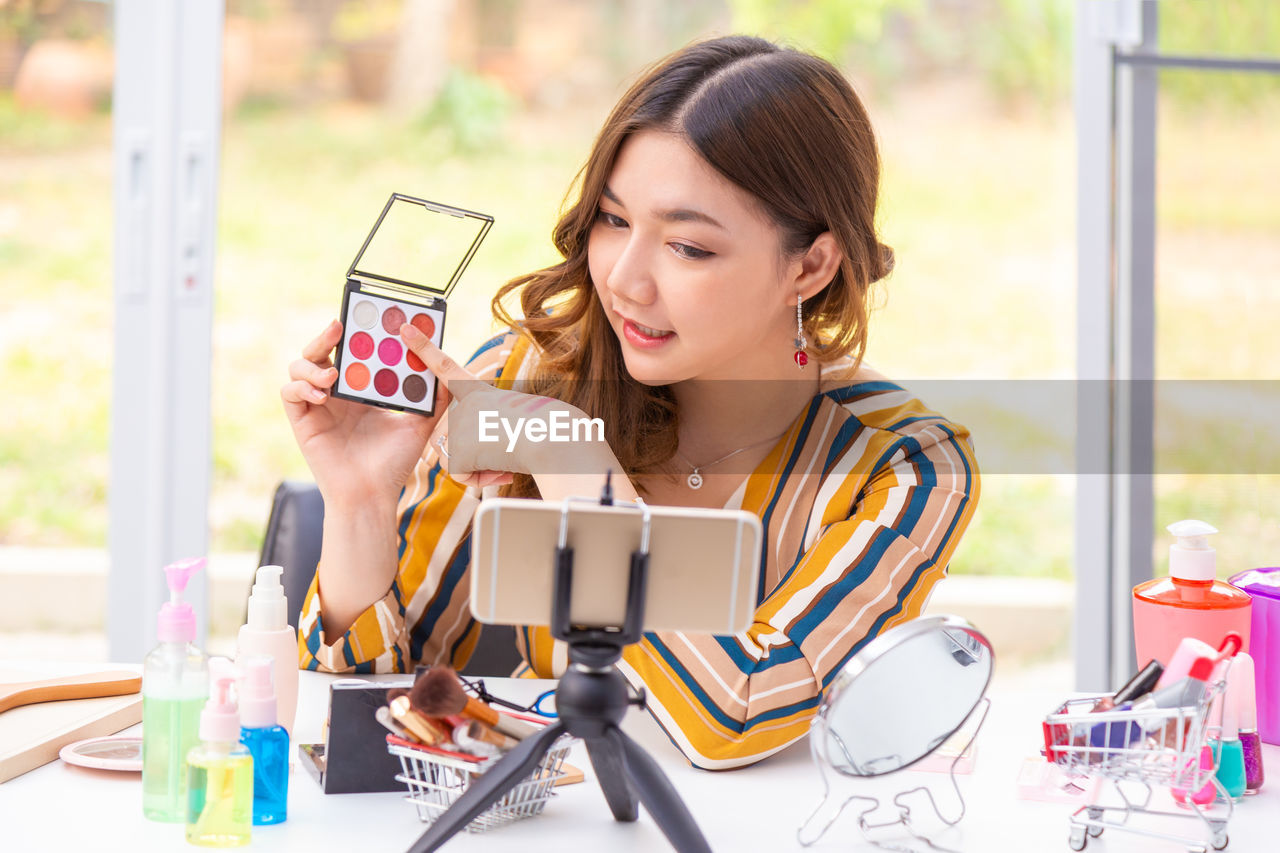 Young woman blogging while holding beauty products on table