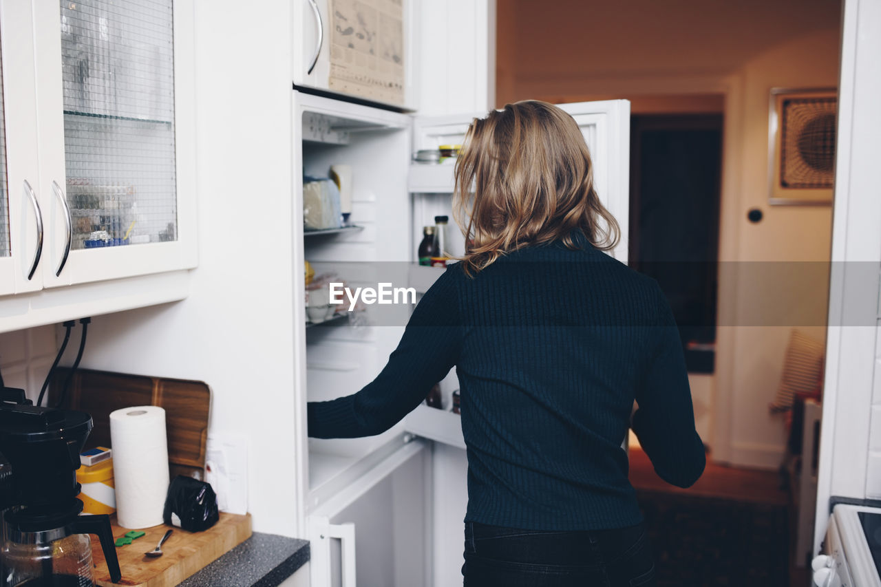 Rear view of woman opening refrigerator in kitchen at home