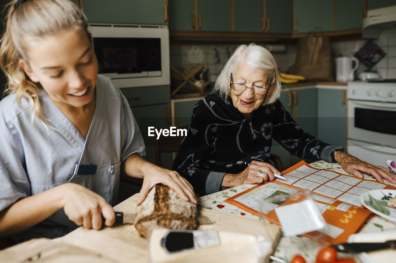 Young female healthcare worker cutting bread loaf sitting by senior woman solving sudoku at home