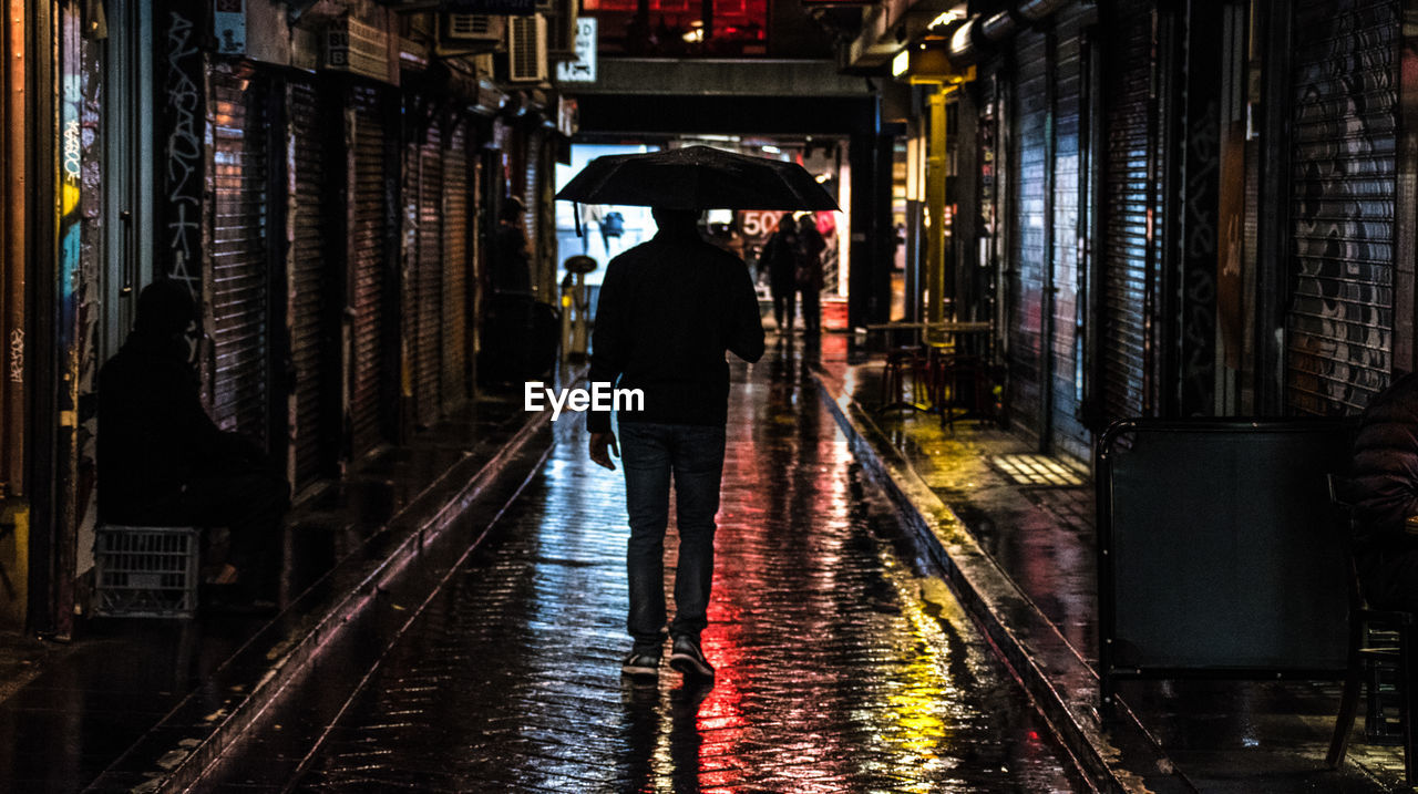 REAR VIEW OF MAN STANDING ON WET ROAD IN CITY