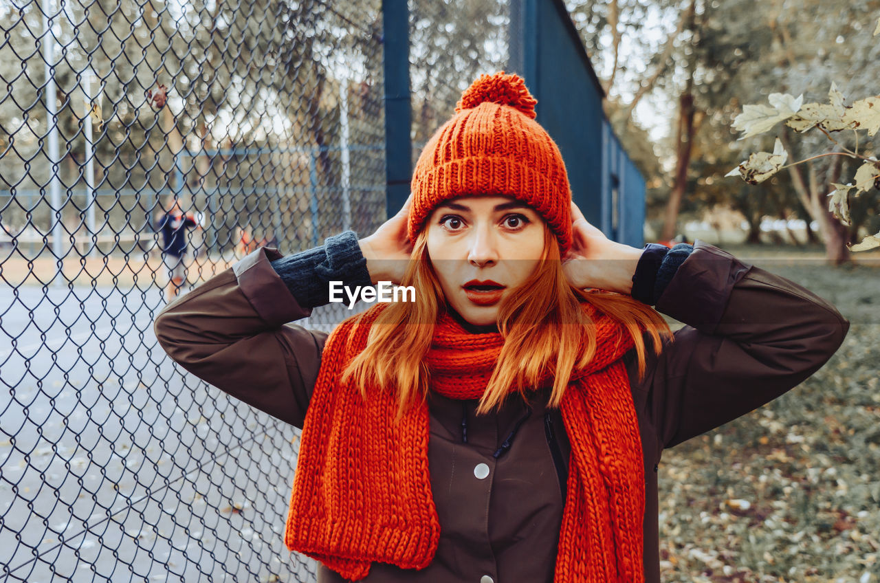 Bright cute young woman in orange warm knitted hat and scarf hands near head. emotion of concern.