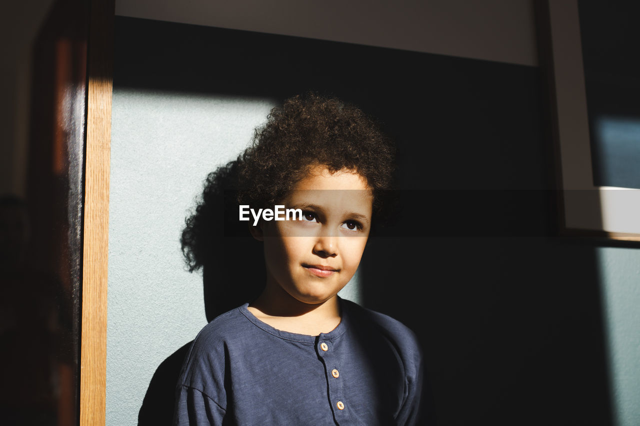 Boy looking away standing in sunlight against wall at home