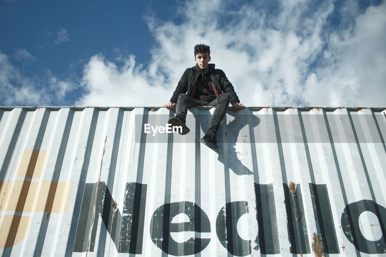 LOW ANGLE VIEW OF YOUNG MAN SITTING ON WALL AGAINST SKY