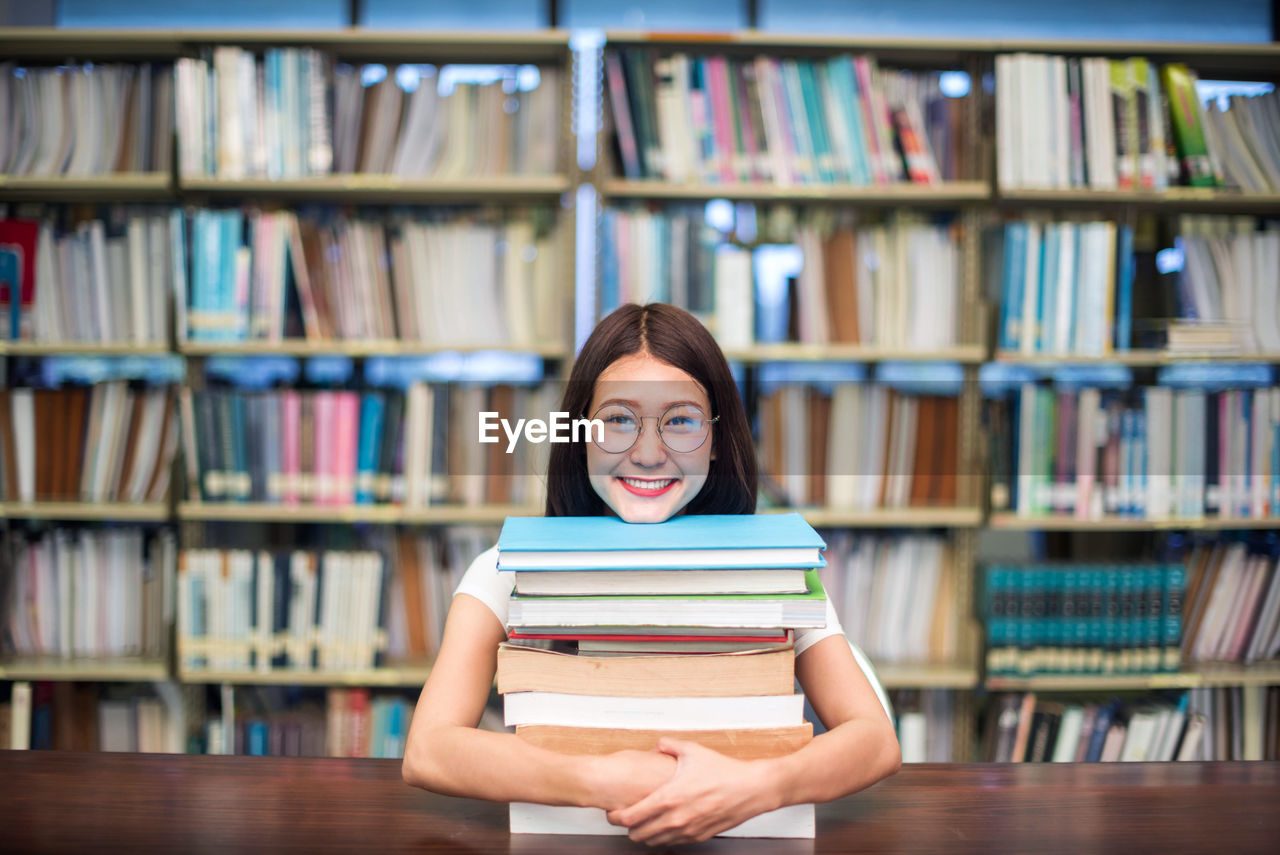 Portrait of smiling female student with stack books on table in library