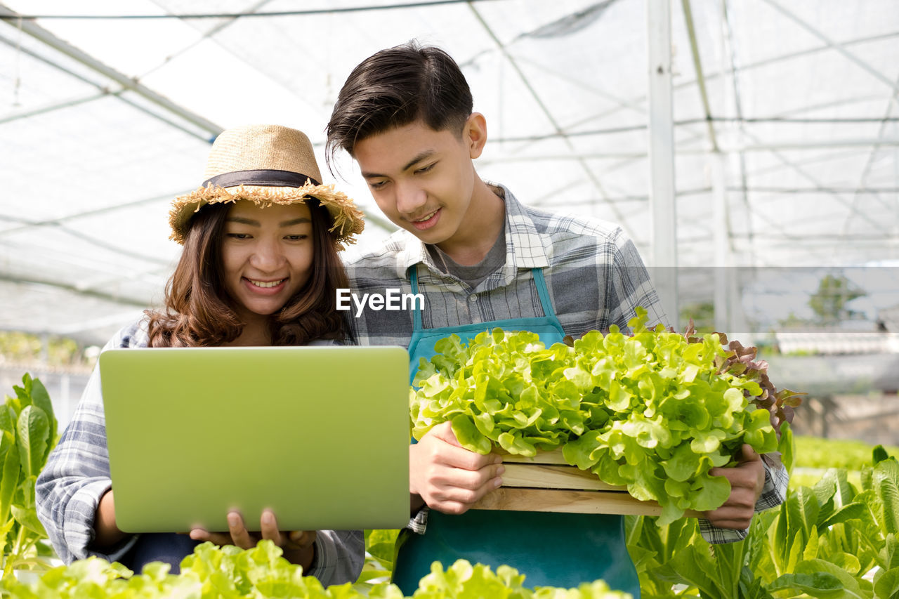 Smiling botanist looking at laptop while holding vegetable at greenhouse