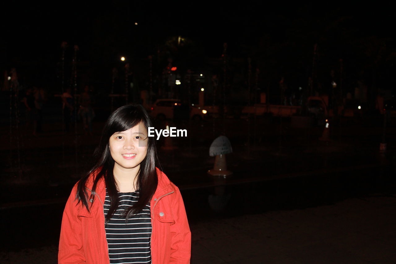 PORTRAIT OF SMILING WOMAN STANDING AT NIGHT