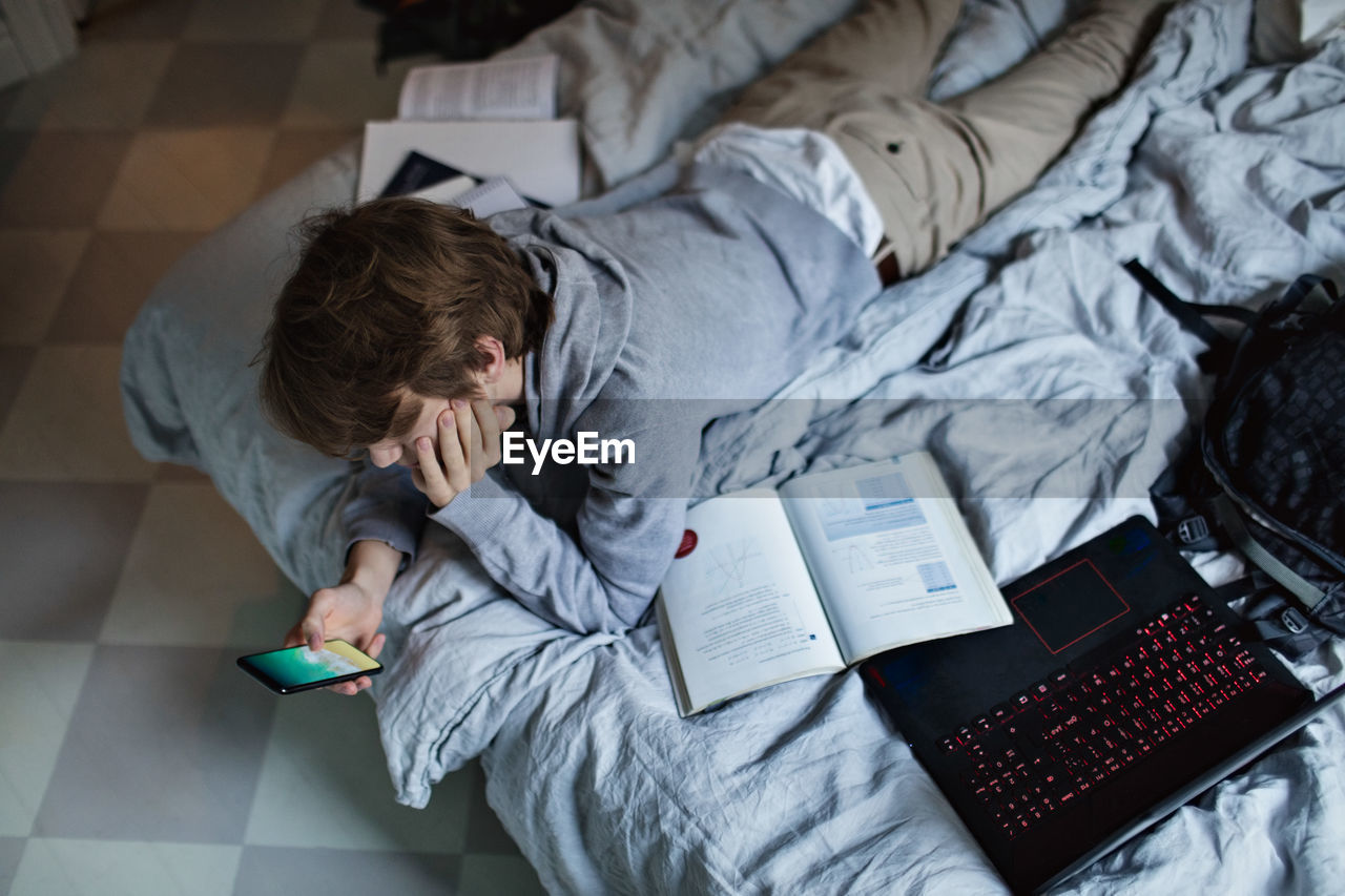 High angle view of teenage boy using mobile phone while lying by books and laptop on bed at home