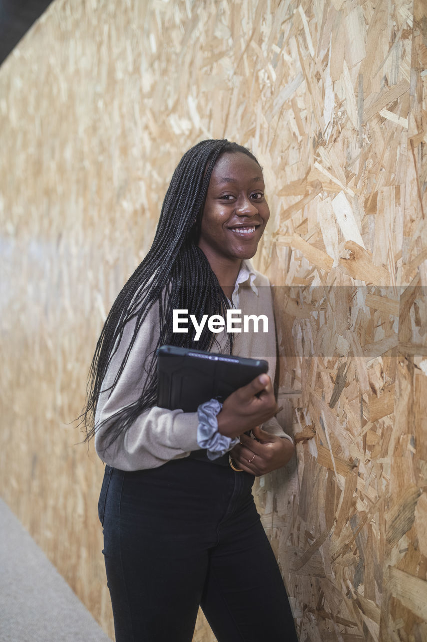 Smiling african american female entrepreneur standing with tablet near wall in coworking space while looking at camera