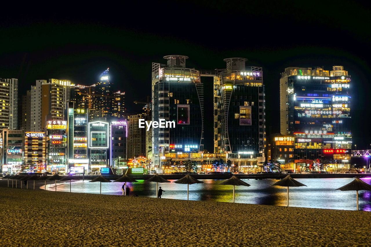 ILLUMINATED BUILDINGS BY RIVER AGAINST SKY AT NIGHT