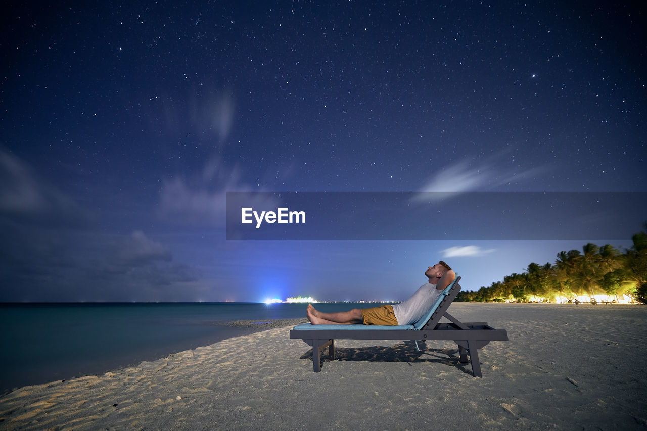 Man lying on sunbed on sand beach and looking up to stars on night sky.