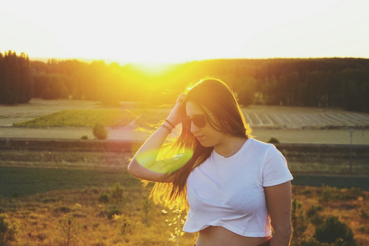 WOMAN STANDING ON FIELD AGAINST SKY AT SUNSET