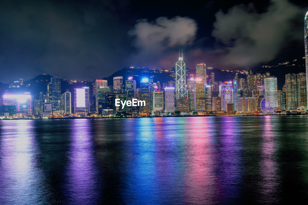 Illuminated buildings by river against sky at night