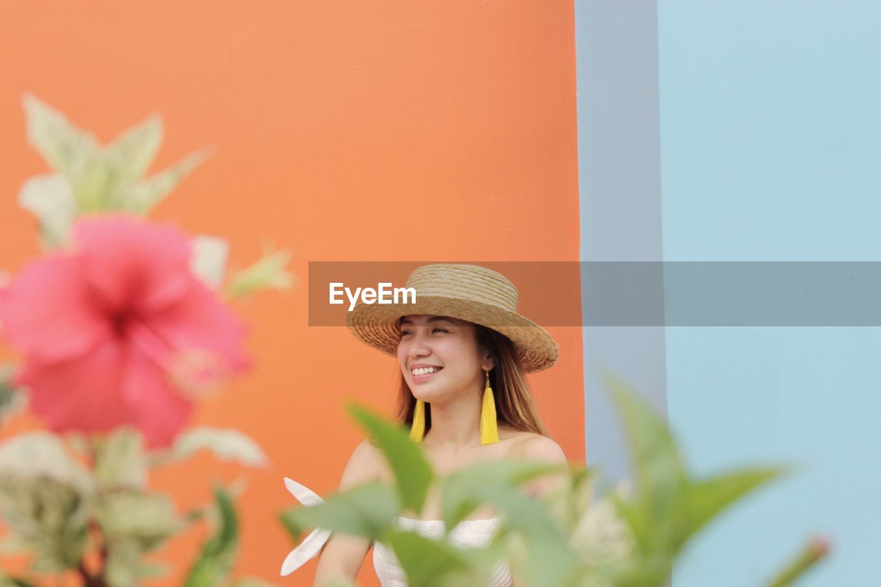 Smiling young woman against wall with plant in foreground