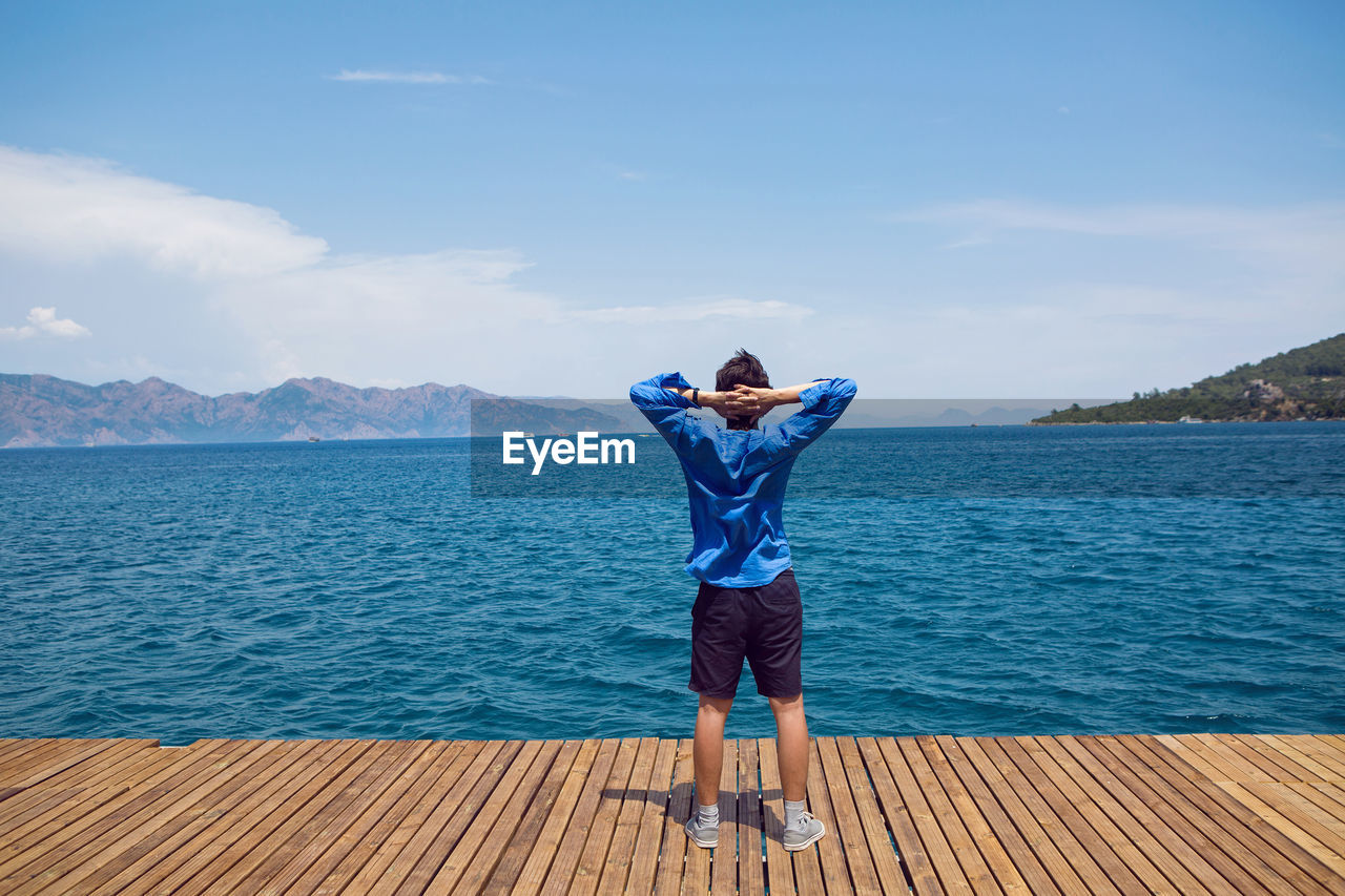 Guy stands rear view by the sea on the pier in a blue shirt in sneakers