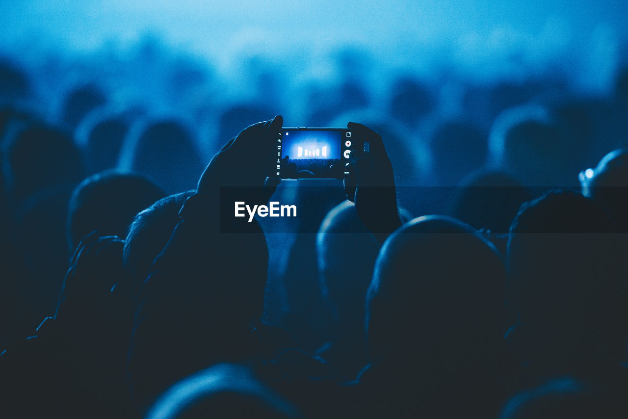 Man in crowd photographing illuminated stage through smart phone at theater