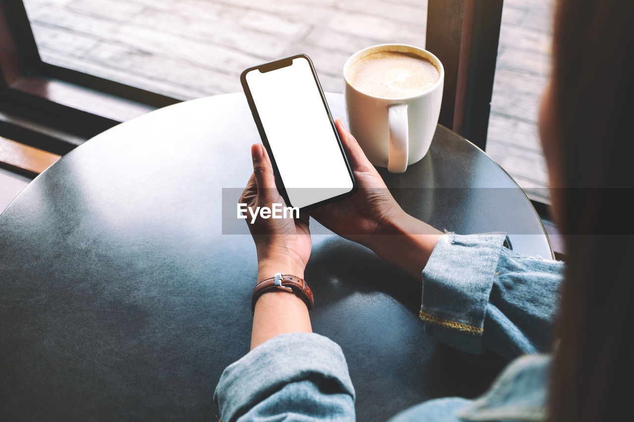 Cropped image of woman using blank phone at table in cafe