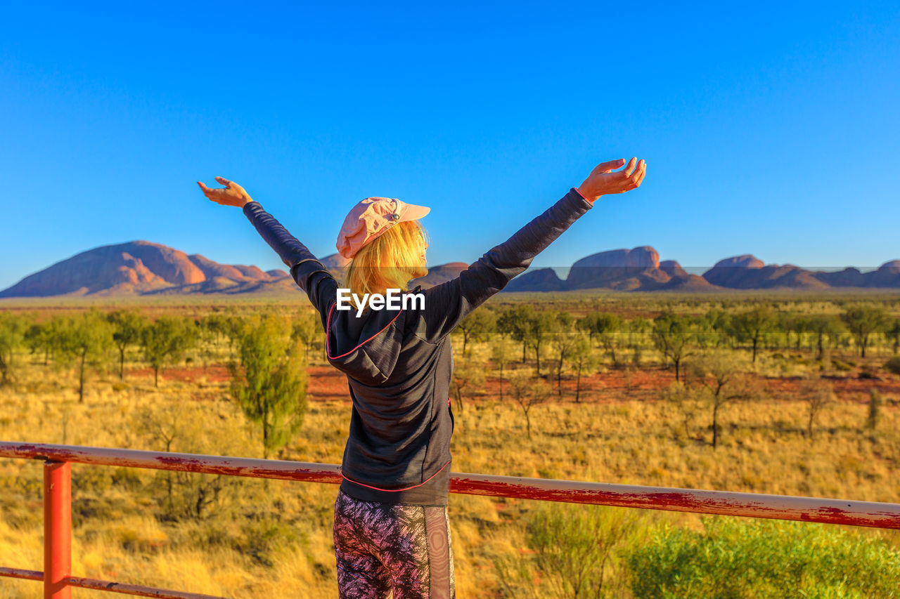 REAR VIEW OF PERSON STANDING ON LANDSCAPE AGAINST BLUE SKY