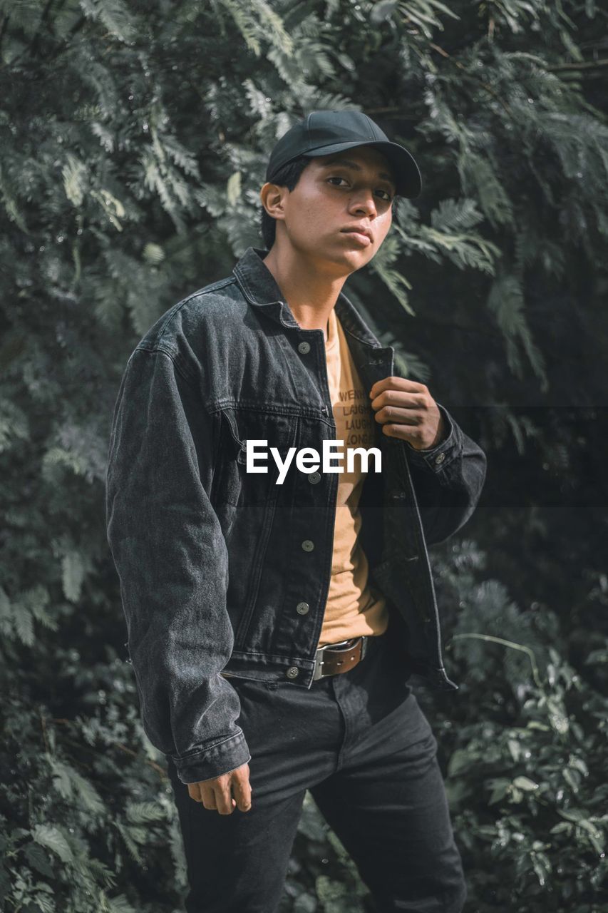 YOUNG MAN LOOKING AWAY WHILE STANDING ON TREE IN FOREST