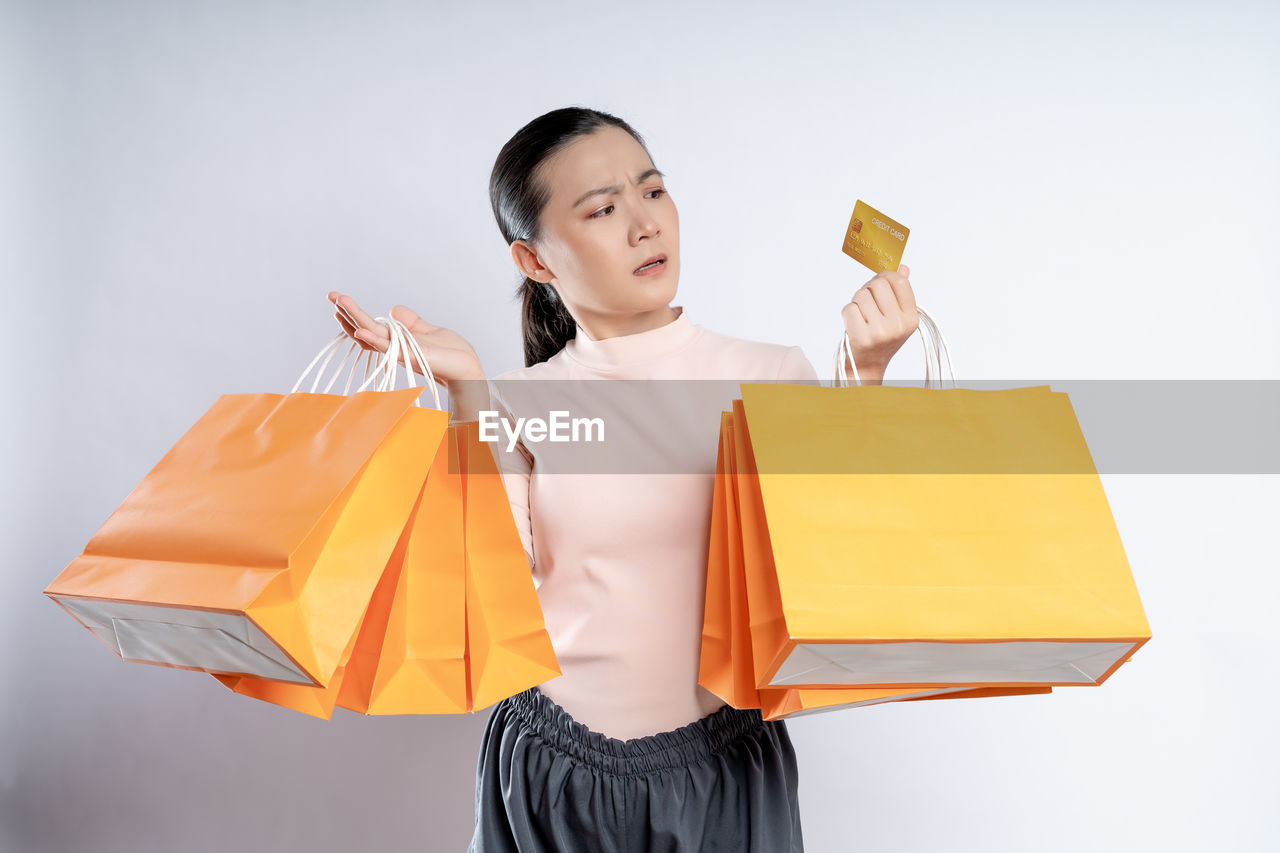 shopping bag, shopping, consumerism, bag, yellow, spending money, retail, orange, happiness, one person, holding, women, paper bag, smiling, studio shot, customer, adult, tote bag, cheerful, emotion, buying, handbag, clothing, carrying, fashion accessory, celebration, casual clothing, indoors, holiday, paper, sale, female, positive emotion, lifestyles, young adult, business, fun, child, gift, copy space, childhood, standing, looking