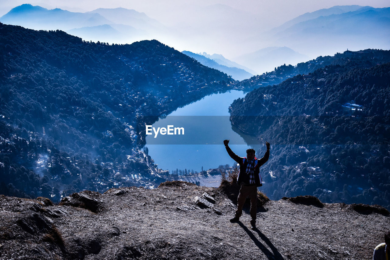 REAR VIEW OF MAN WITH ARMS RAISED AGAINST MOUNTAIN RANGE