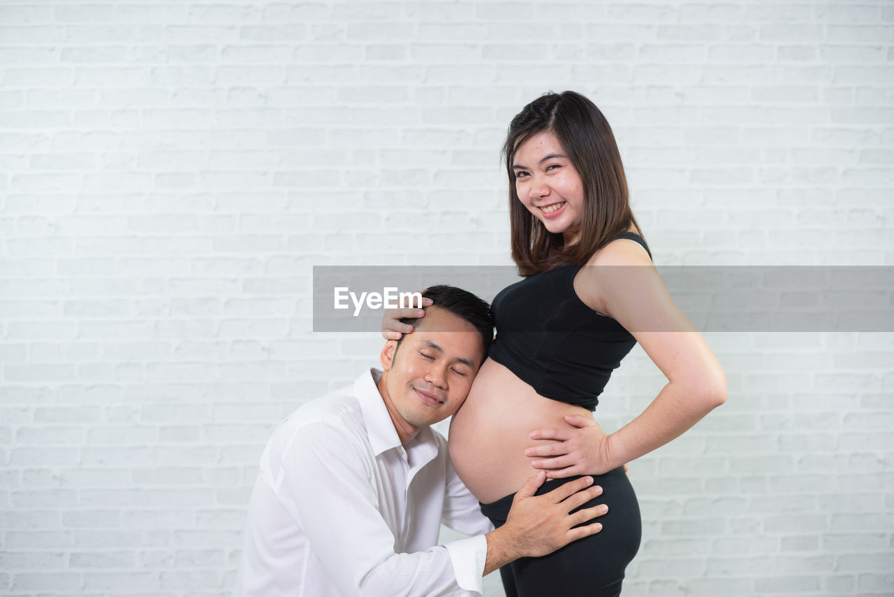 Mid adult man with eyes closed listening to pregnant woman belly against wall