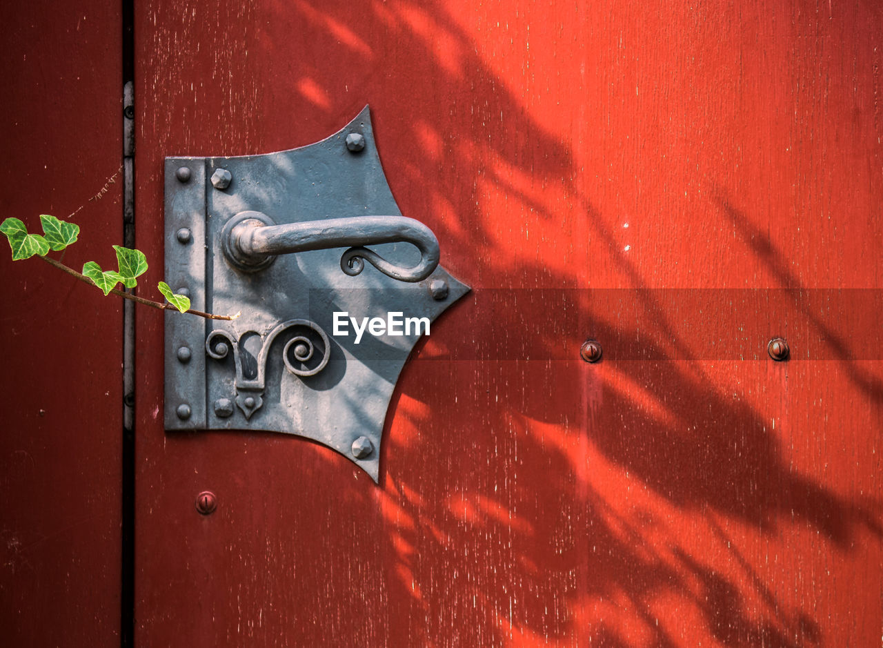 An old red door with a vintage knob and a modern door lock. a plant is growing in front of it.