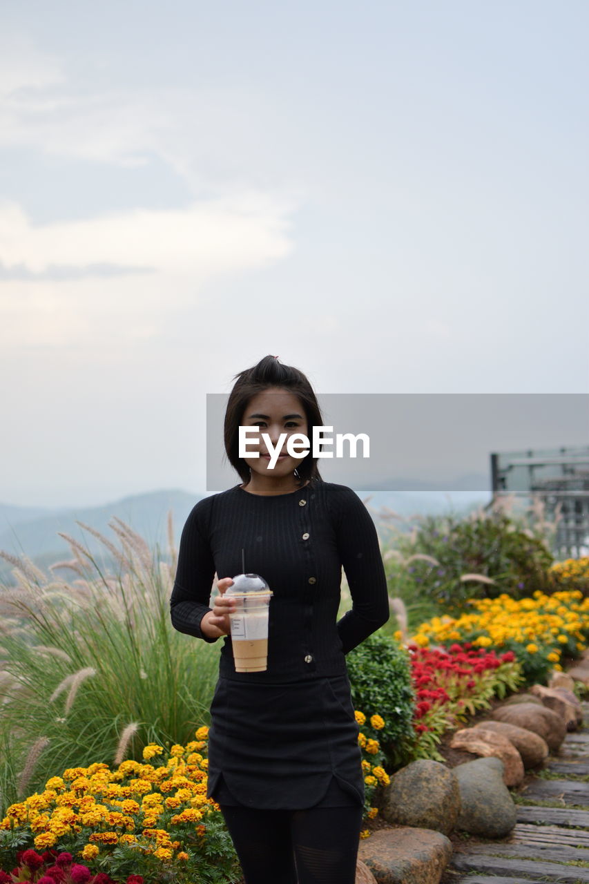 Portrait of smiling young woman holding coffee while standing on grassy field against sky