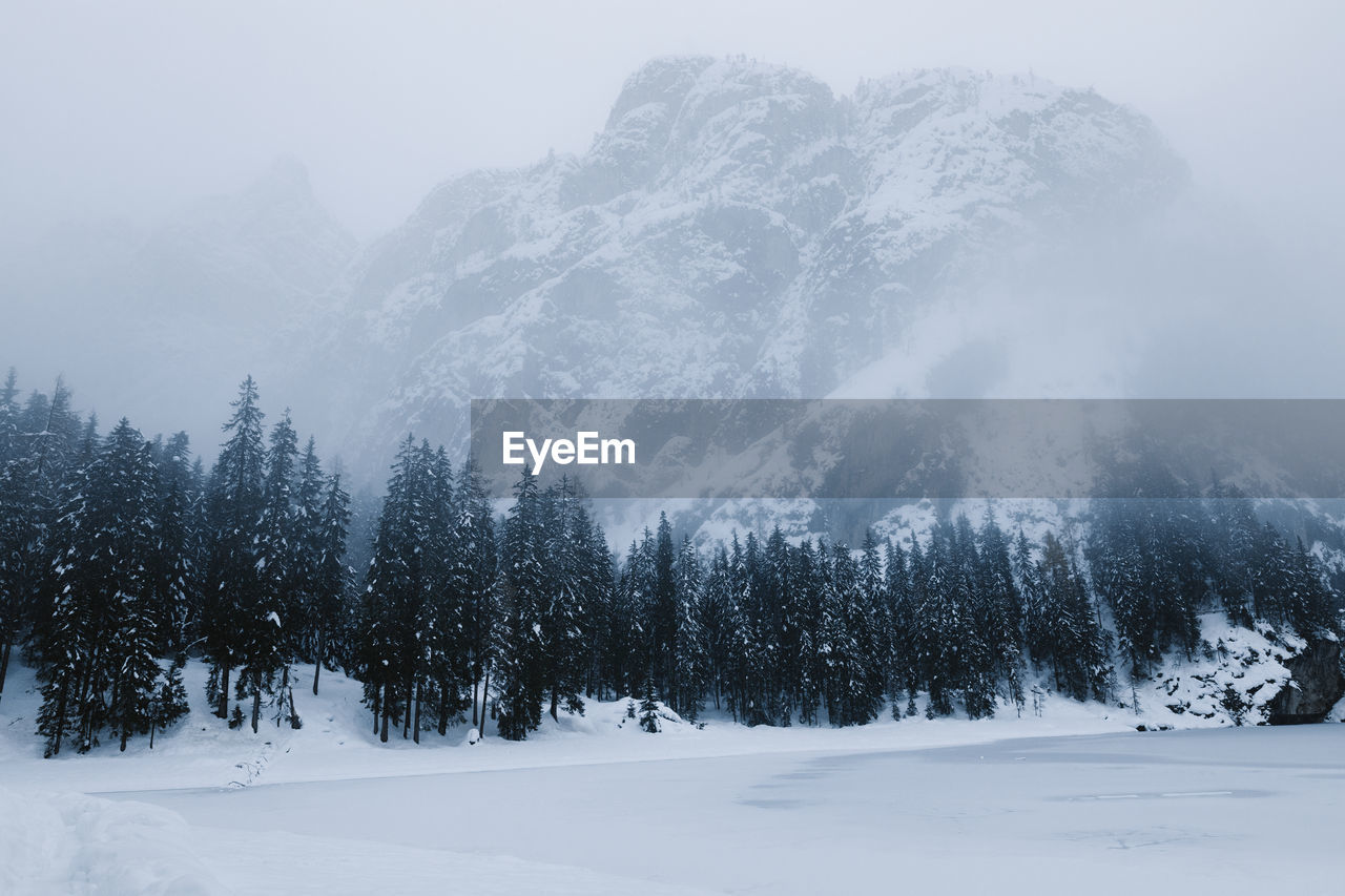 Severe scenery of frozen river surrounded by coniferous woods and rough rocky mountains covered with snow during snowfall in cold winter day