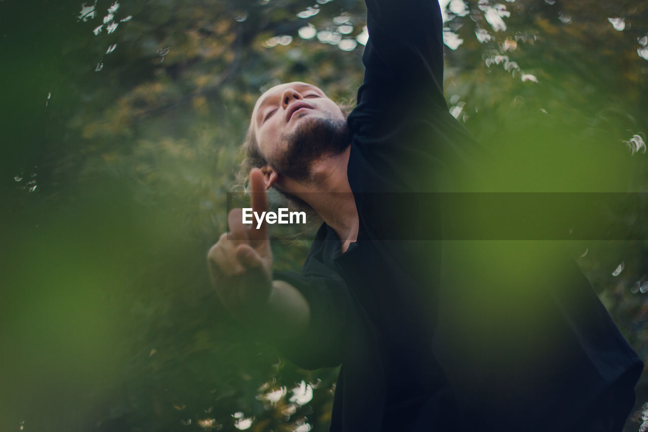 Thoughtful young man dancing with eyes closed against trees 