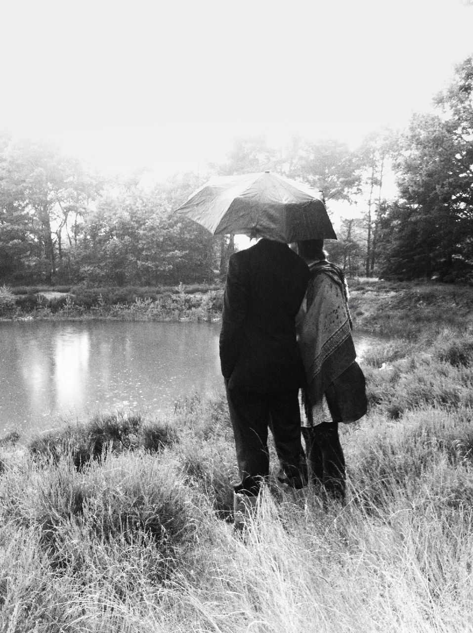 Man and woman under umbrella on grassy field by pond