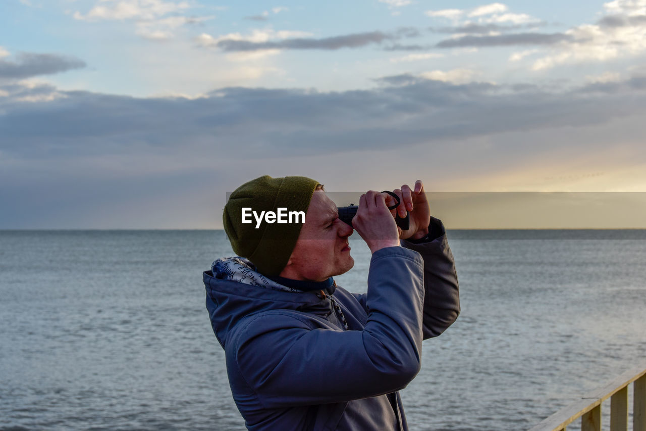 Profile view of young man looking through binoculars against sea during sunset
